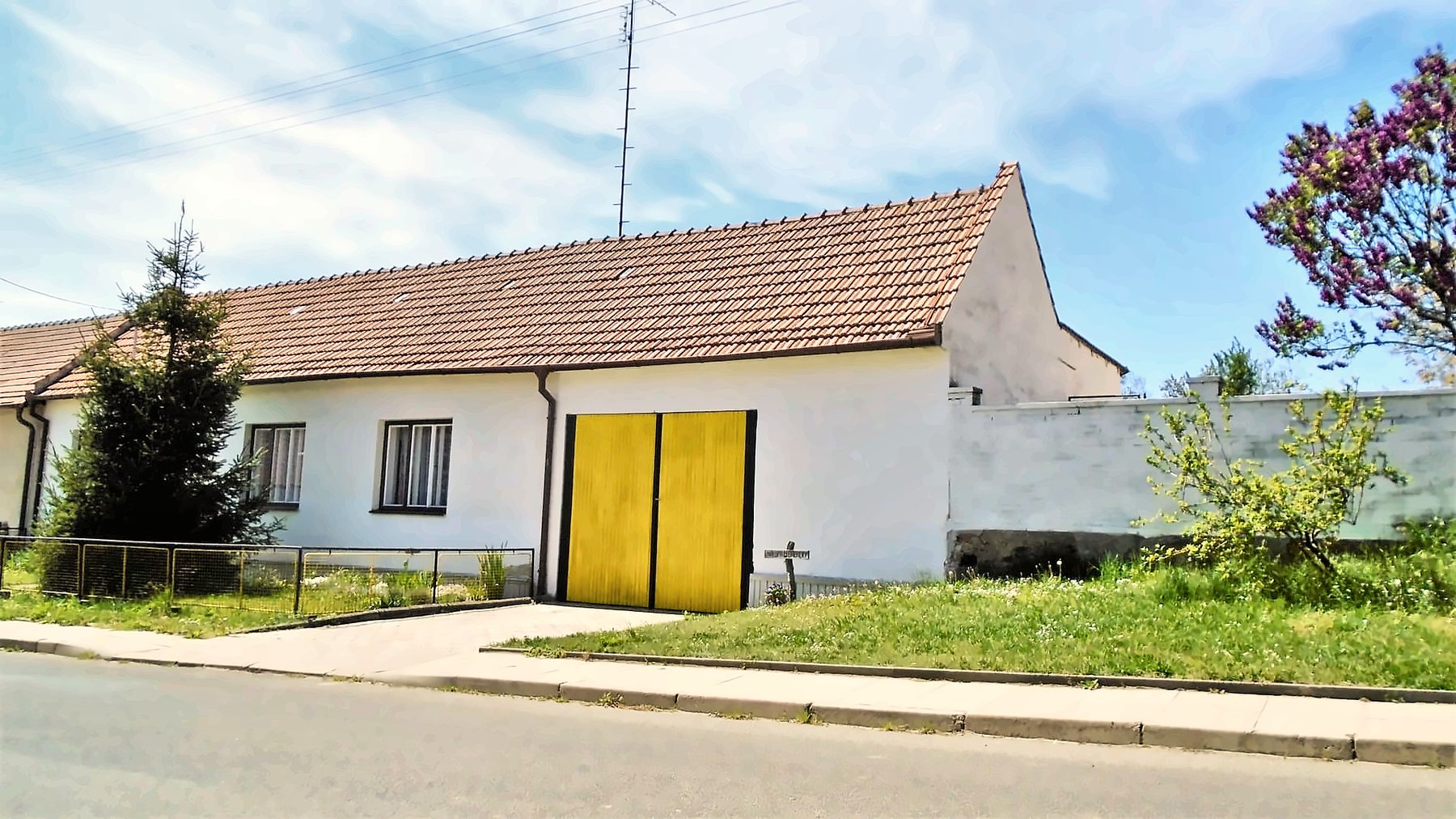 Dlouhá bílá chaloupka s červenou střechou a žlutými vraty u silnice